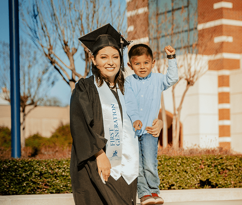 Mom in graduation cap and gown stands with her toddler son.