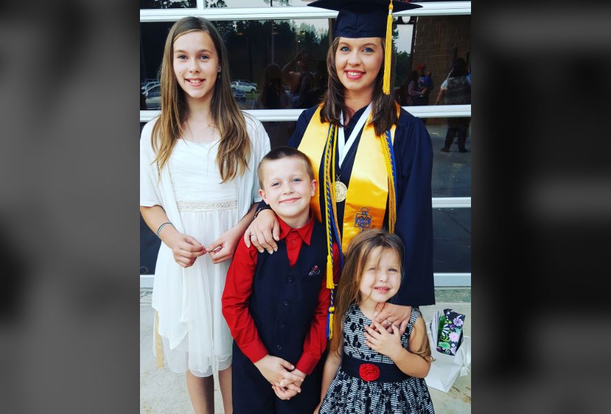 Mom in Graduation garb stands with three kids.