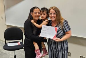 A mom holds her toddler giril while another woman holds a certificate. 