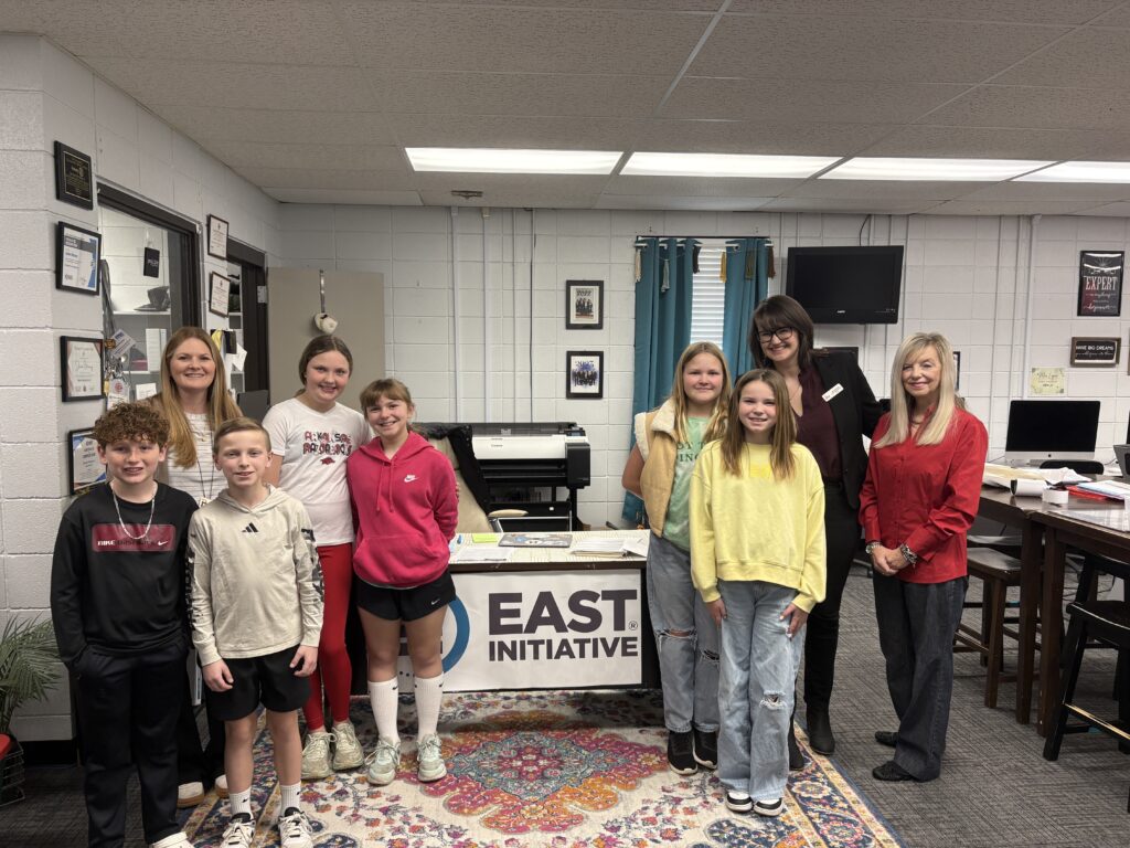 Fifth graders and adults stand in a classroom with an EAST Initative sign.