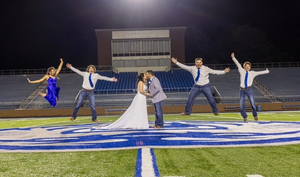 Groom and bride kiss on a football field while their four kids jump in the air.