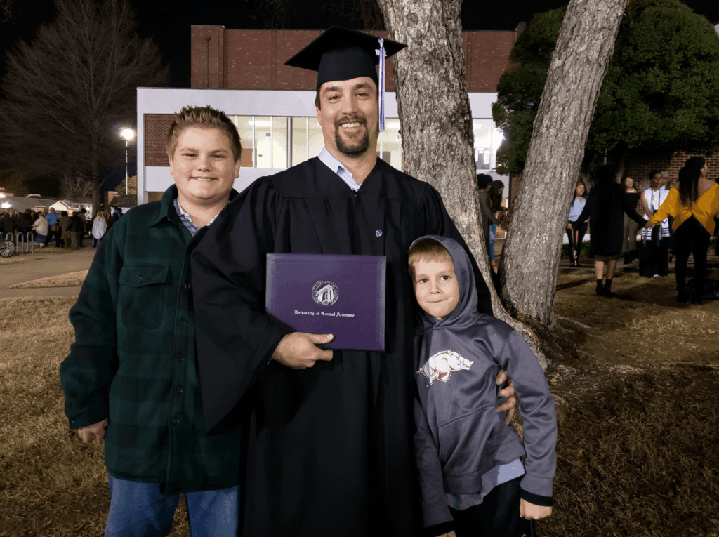 CJ Summerville, dressed in graduation cap and gown, holds his UCA diploma. His two sons are on either side of him, and they're all standing in front of a tree.