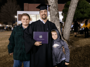 CJ Summerville, dressed in graduation cap and gown, holds his UCA diploma. His two sons are on either side of him, and they're all standing in front of a tree. 