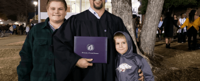 CJ Summerville, dressed in graduation cap and gown, holds his UCA diploma. His two sons are on either side of him, and they're all standing in front of a tree.