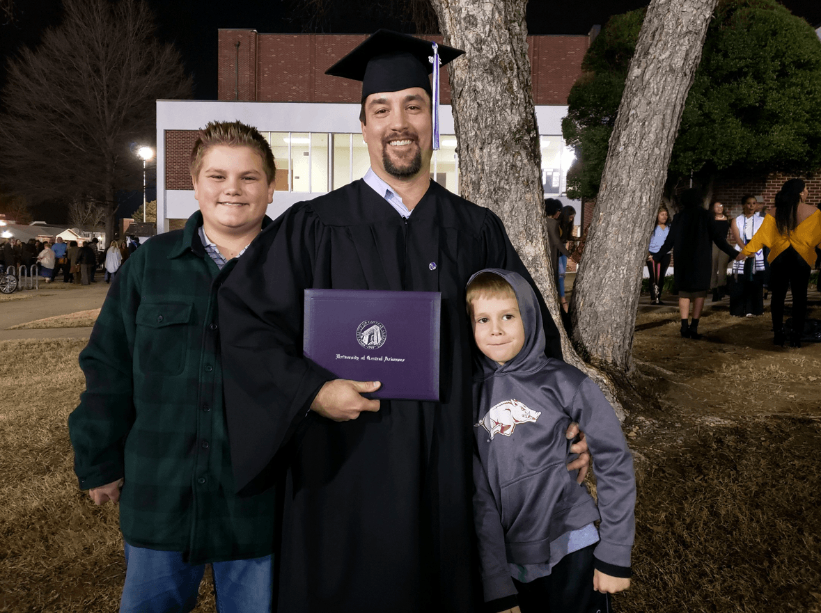 CJ Summerville, dressed in graduation cap and gown, holds his UCA diploma. His two sons are on either side of him, and they're all standing in front of a tree.
