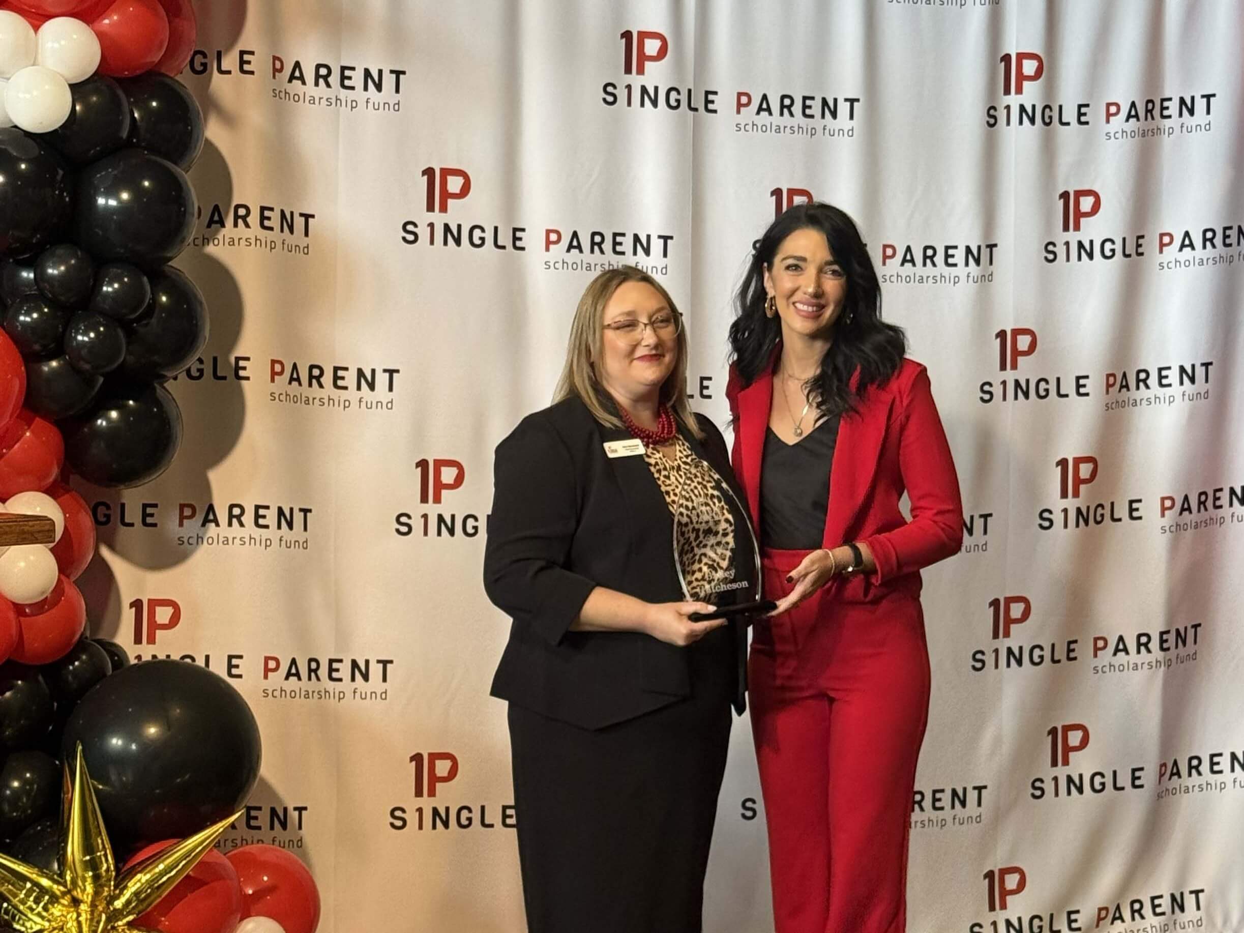 Bailey Hutcheson right-Distinguished Alumna Jenn Morehead and Bailey Hutcheson hold an award in front of a step-and-repeat